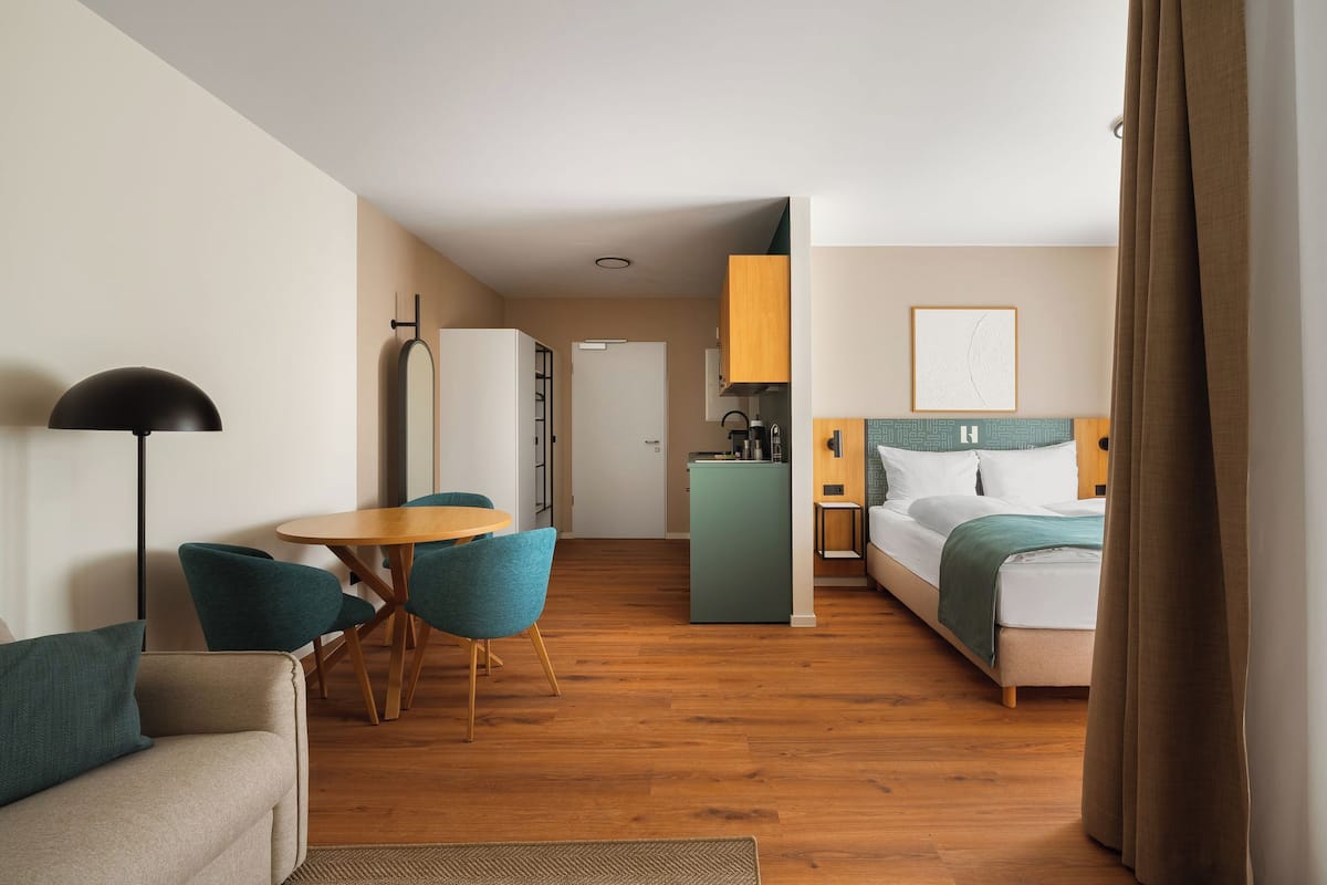 A hotel room with a bed, a desk, a chair, a lamp, and a kitchenette.