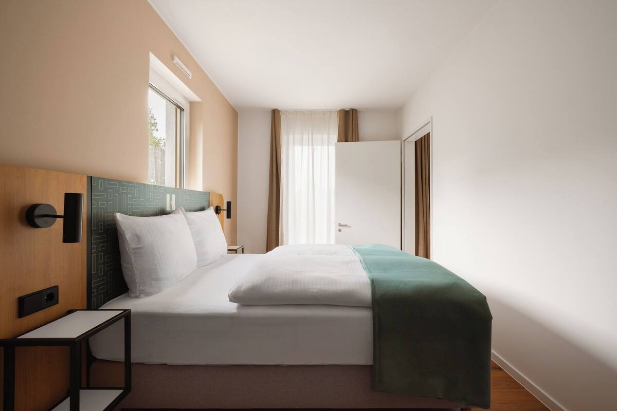 A modern hotel room with a neatly made bed, a bedside table, a wall-mounted lamp, and a window with curtains.
