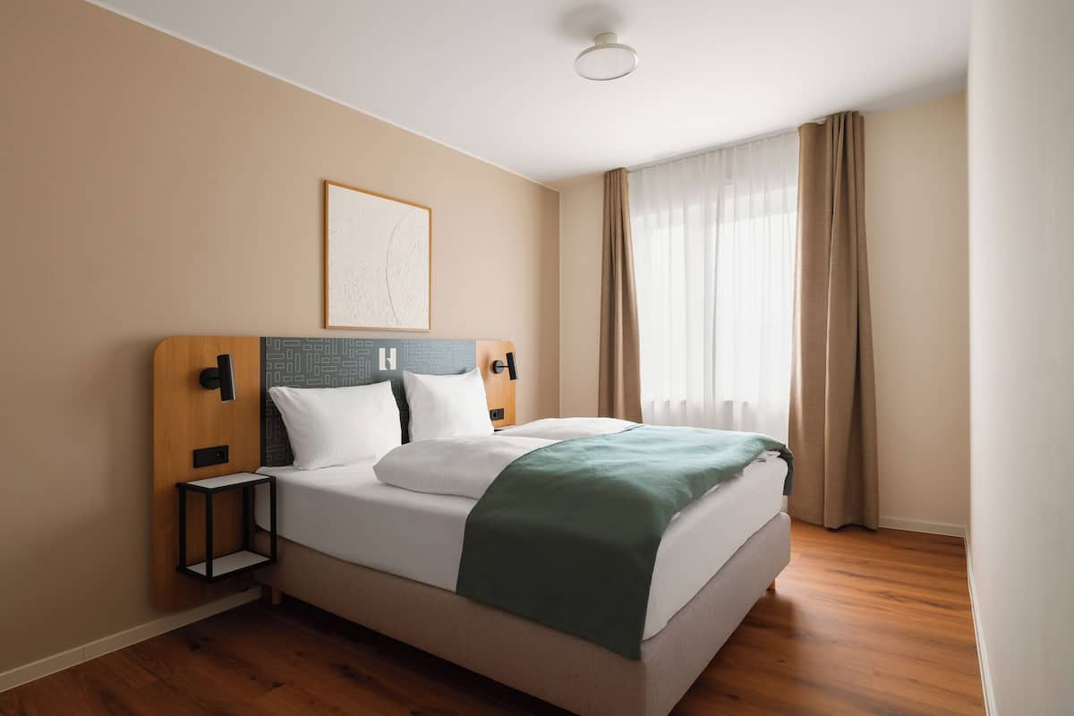 A hotel room with a bed, a bedside table, a wall-mounted artwork, and a window with curtains.