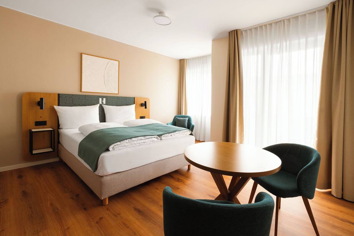 A hotel room with a bed, a bedside table, a chair, a round table, and a window with curtains.