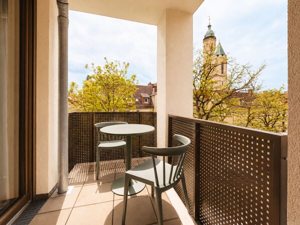 Un balcón con mesa y sillas, vista de un edificio con torre y árboles de hojas amarillas.