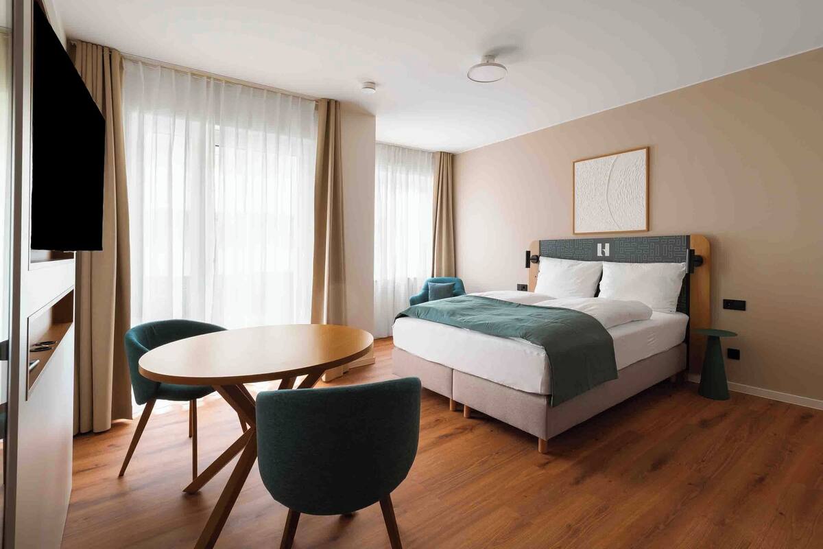 A hotel room with a bed, a round table, two chairs, a TV, and a painting on the wall.