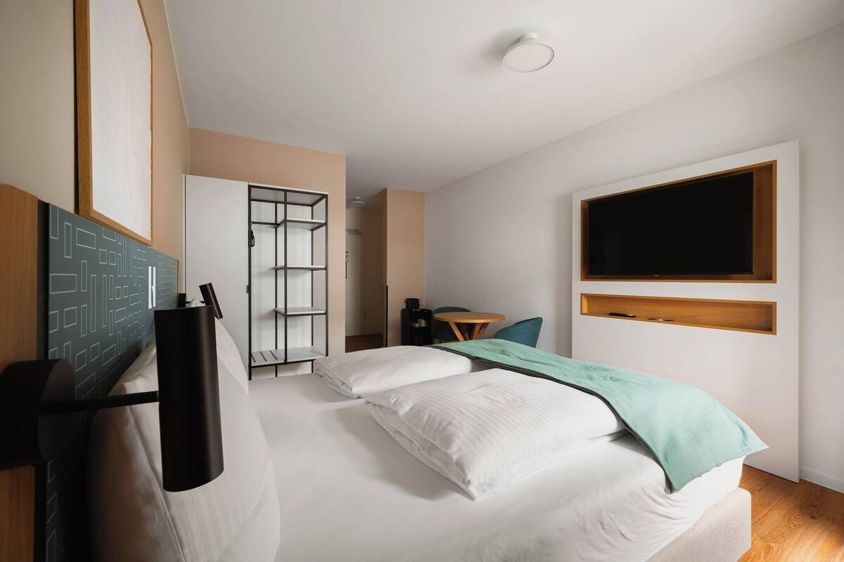 A modern hotel room with a bed, a desk, a chair, a TV, and a wardrobe.