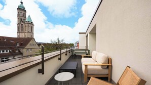 Ein Balkon mit Blick auf einen Kirchturm, eine Couch, ein kleiner Tisch und ein Korbstuhl.