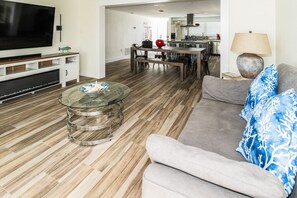 A modern living room with a glass coffee table, a grey sofa with patterned cushions, a flat-screen TV, and a dining area with a wooden table and chairs. - Mejia's Little House - Cozy Waterfront Escape (New Port Richey)