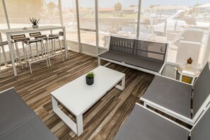 A modern outdoor seating area with a white table, grey cushioned benches, and a view of a marina. - Mejia's Little House - Cozy Waterfront Escape (New Port Richey)
