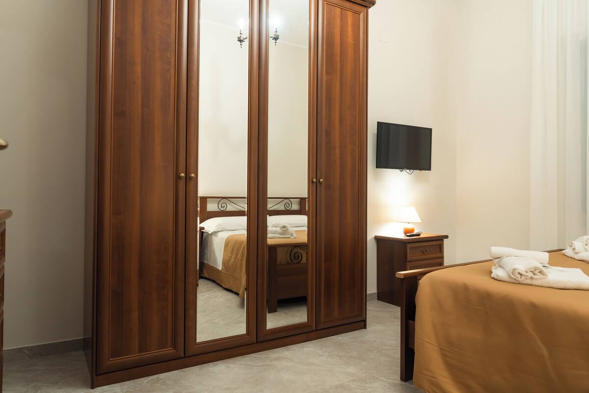 A bedroom with a wooden wardrobe, a bed with a beige bedspread, a bedside table with a lamp, and a flat-screen TV mounted on the wall.