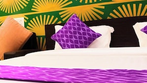 Een hotelkamer met een zonburstpatroon in groen en geel op de muur, een bed met witte en paarse kussens en een paars plaid.