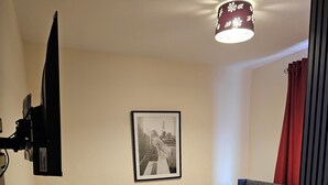 Une chambre à coucher avec un lit, un ventilateur sur pied, un tableau encadré accroché au mur et un téléviseur fixé au mur.