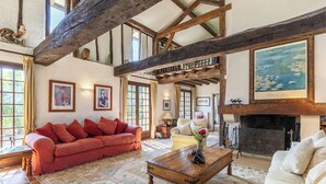 A spacious living room with a high ceiling, exposed wooden beams, a fireplace, and a large window. - Tarrit de Bas with gated pool and total privacy (Castelnau d’Auzan Labarrère)