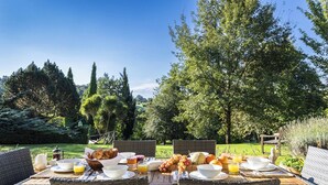 An outdoor dining table set with breakfast items, wicker chairs, and a view of a lush garden. - Tarrit de Bas with gated pool and total privacy (Castelnau d’Auzan Labarrère)