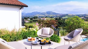 A patio with wicker chairs, a table with food and drinks, and a view of rolling hills and distant houses. - Villa Souraïde with pool, games room and Basque views (Souraide)