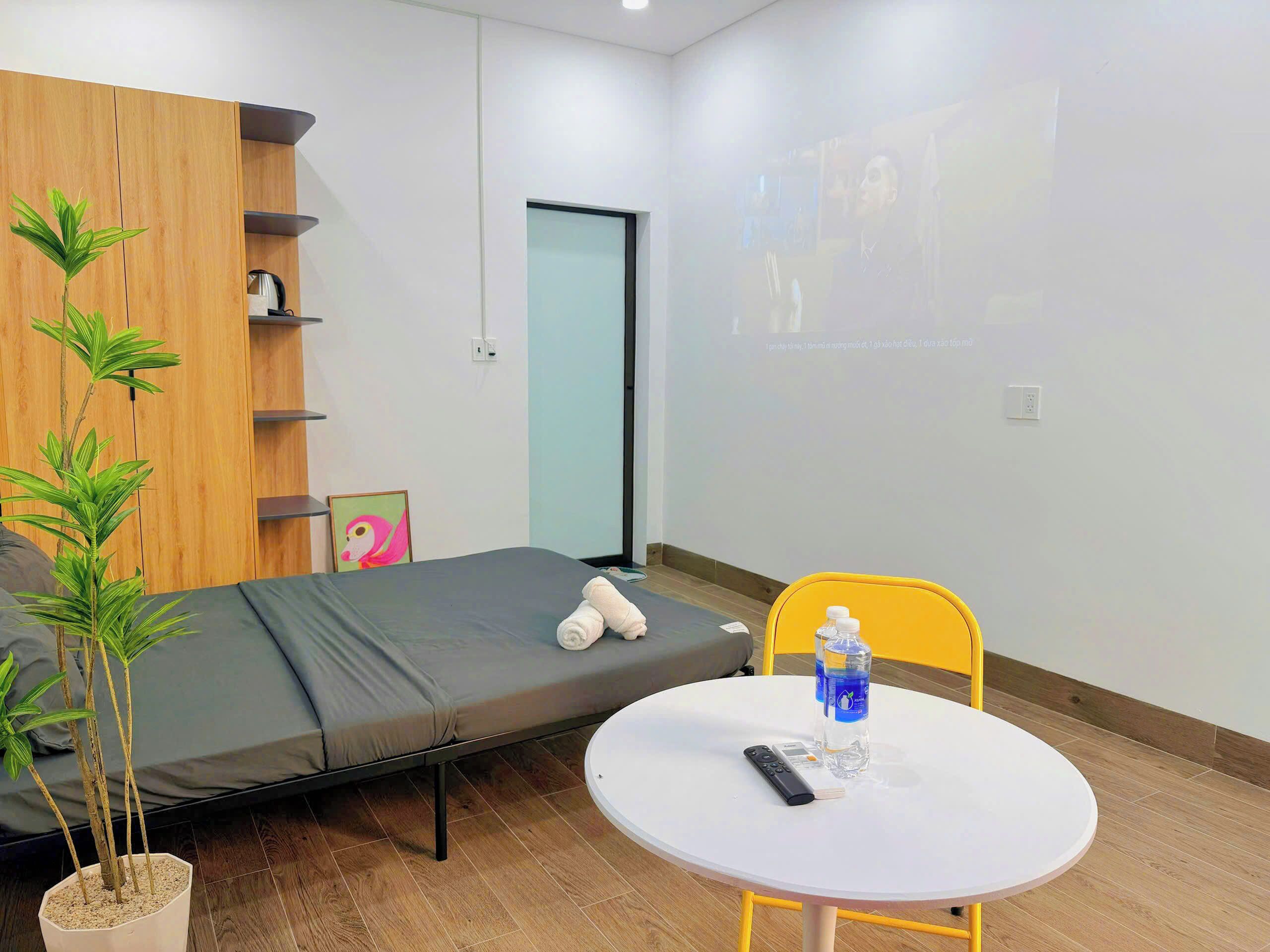 A room with a bed, a small table with a remote control, a water bottle, and a potted plant.
