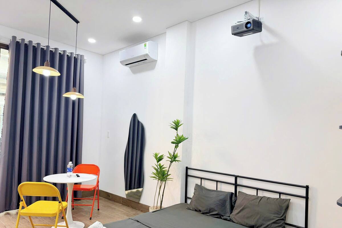 Quarto de hotel com uma cama, uma escrivaninha com cadeira, um aparelho de ar condicionado, um projetor e um vaso de planta.