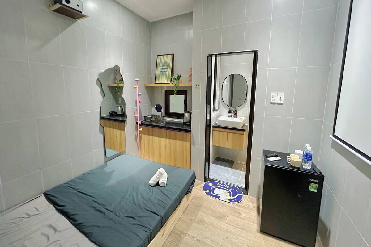 Quarto de hotel moderno com cama, pequena copa, espelho e mini-geladeira.
