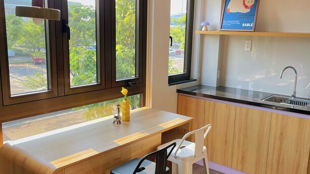 A modern kitchen with wooden cabinetry, a white countertop, a sink, and a window with a view of greenery outside. - Corgi House Da Nang- Airport City Center (Da Nang)