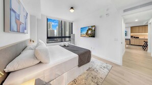Een moderne hotelkamer met een bed, een groot raam met uitzicht over de stad, een flatscreen televisie en een keukenruimte zichtbaar door een geopende deur.