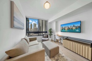 A modern living room with a sofa, a TV, a desk, and a view of a cityscape through large windows. - World Cup 2026 · 2BR Walk to FIFA Fan Fest · Pool (Miami)