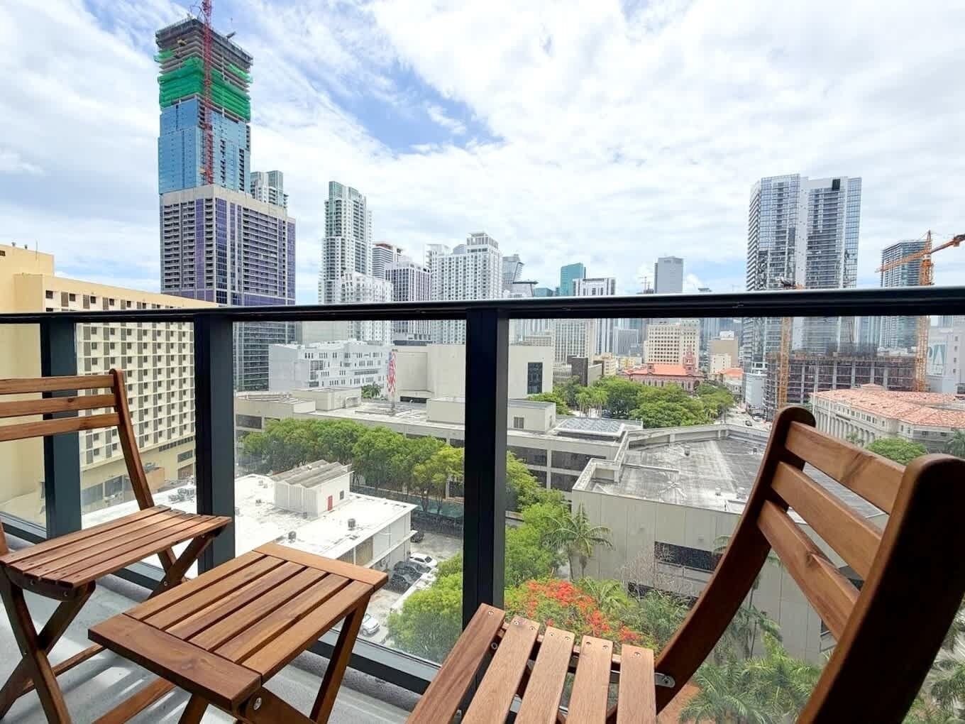Un balcón con dos sillas de madera y una mesa, con vistas a un paisaje urbano que incluye varios edificios y una grúa de construcción.