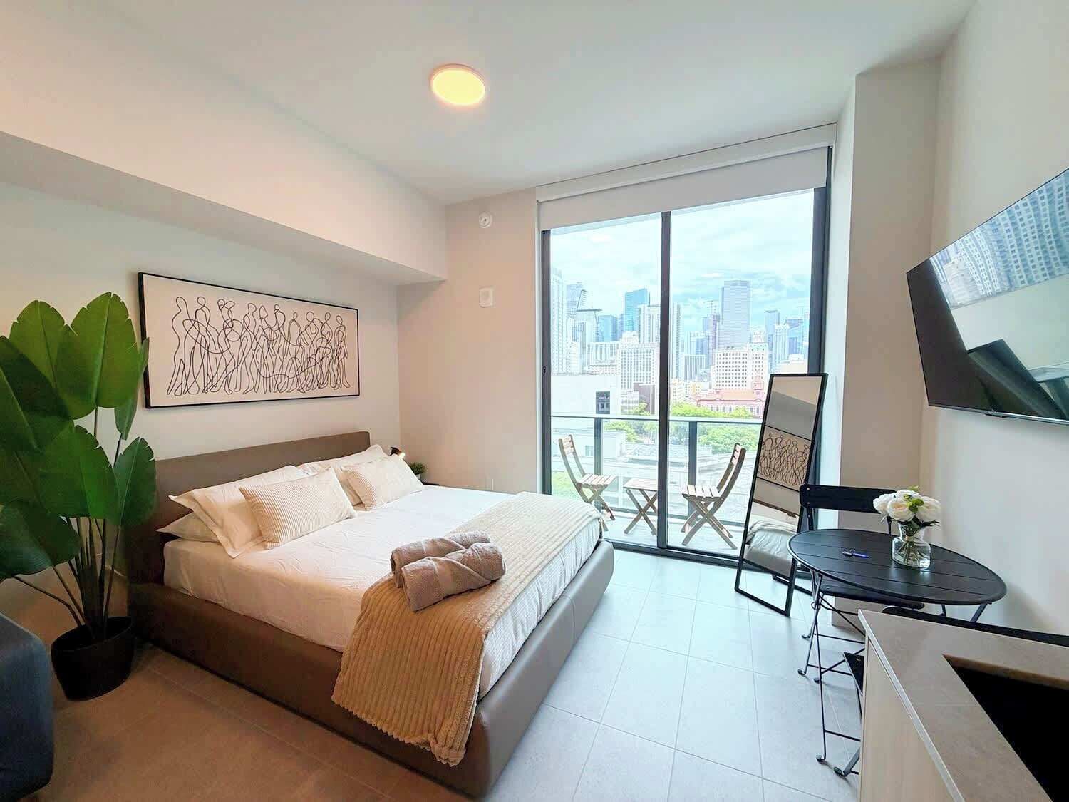 Ein modernes Hotelzimmer mit Bett, einem Balkon mit Blick auf die Stadt, einem kleinen Tisch und einem Blumentopf.