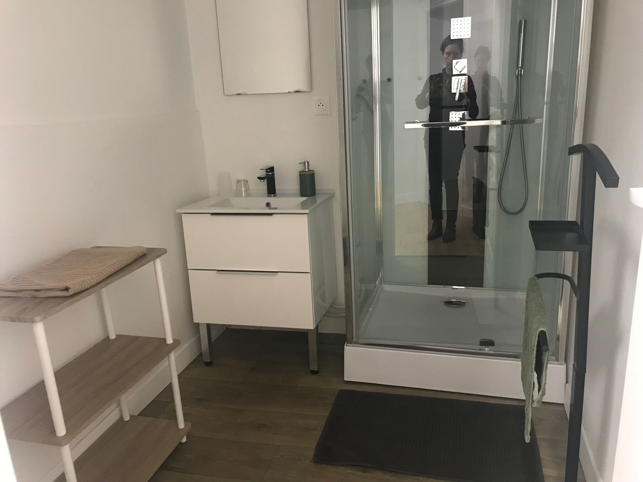 A bathroom with a glass shower enclosure, a sink with a mirror, and a small wooden shelf.