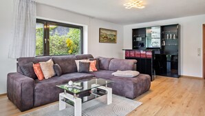 A modern living room with a grey sofa, a glass coffee table, and a kitchen area with cabinets and a refrigerator.