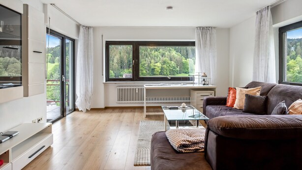 A modern living room with a large window, a sofa, a glass coffee table, and a white desk.