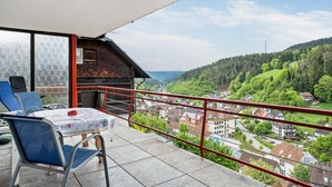 A balcony with a table and chairs, a red railing, and a view of a village and forested hills.