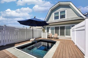 Ein Schwimmbad mit Holzterrasse, weißem Zaun und einem Haus mit Satteldach und großen Fenstern.