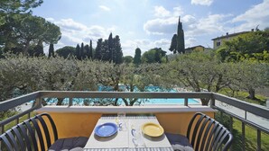 Un balcón con una mesa lista para dos personas, con vista a una piscina y un jardín con árboles.