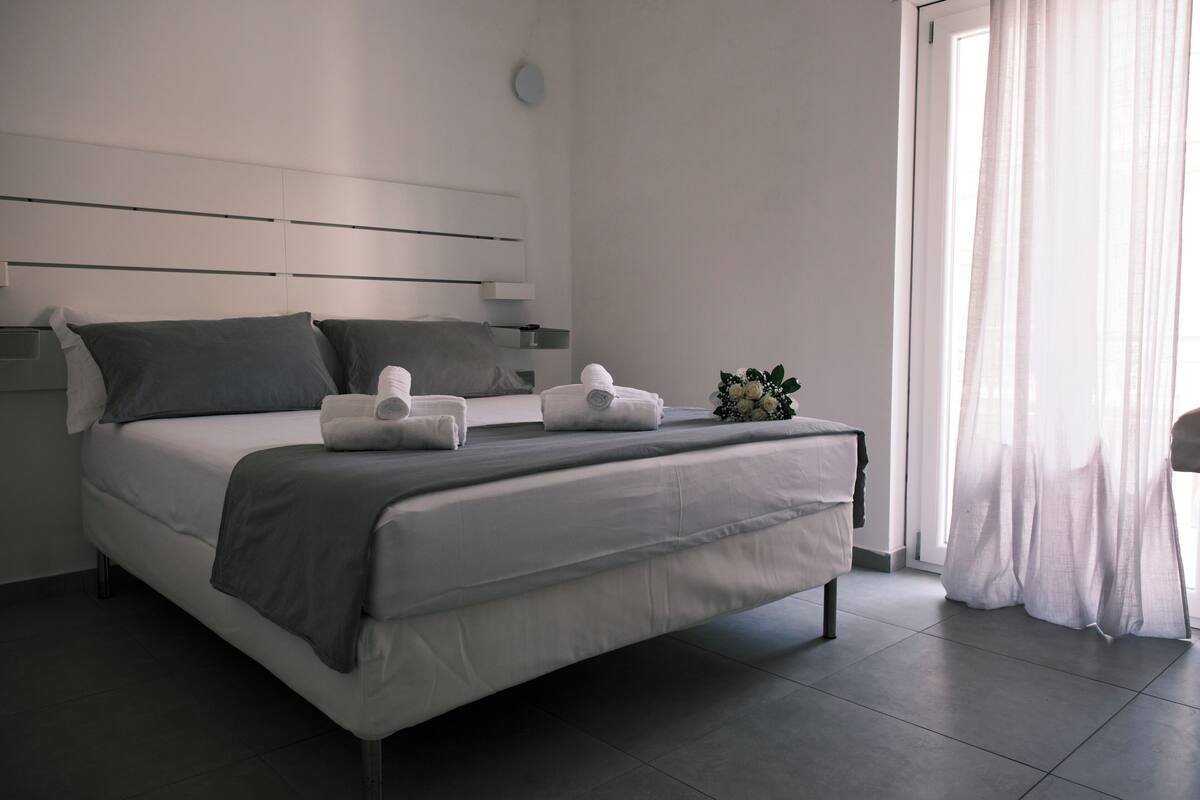 Ein modernes Schlafzimmer mit Bett, Kissen, Nachttisch, Fenster mit Vorhängen und einer kleinen Pflanze.