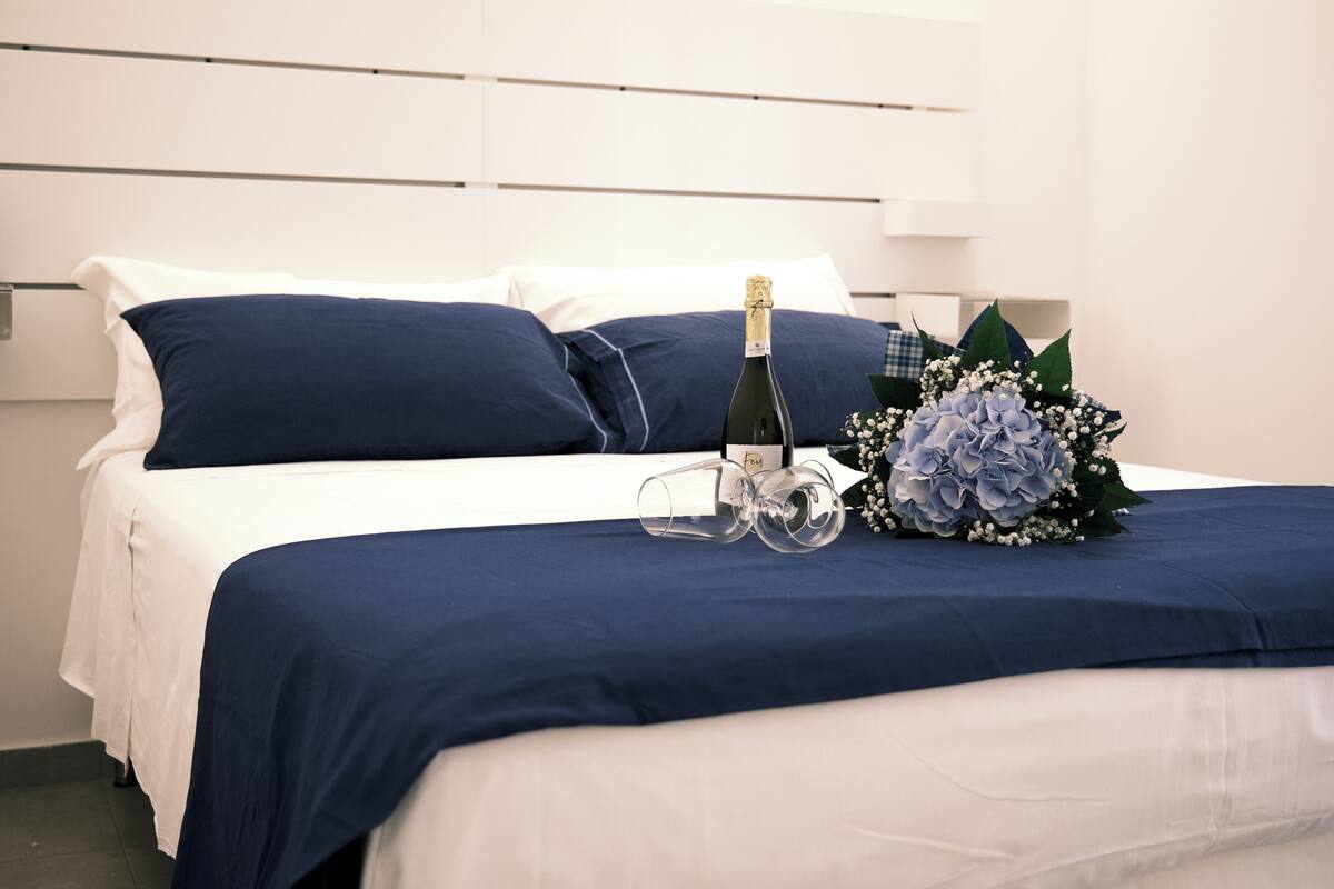 Ein ordentlich bezogenes Bett mit einer blau-weißen Tagesdecke, eine Champagnerflasche und eine Blumenkomposition.