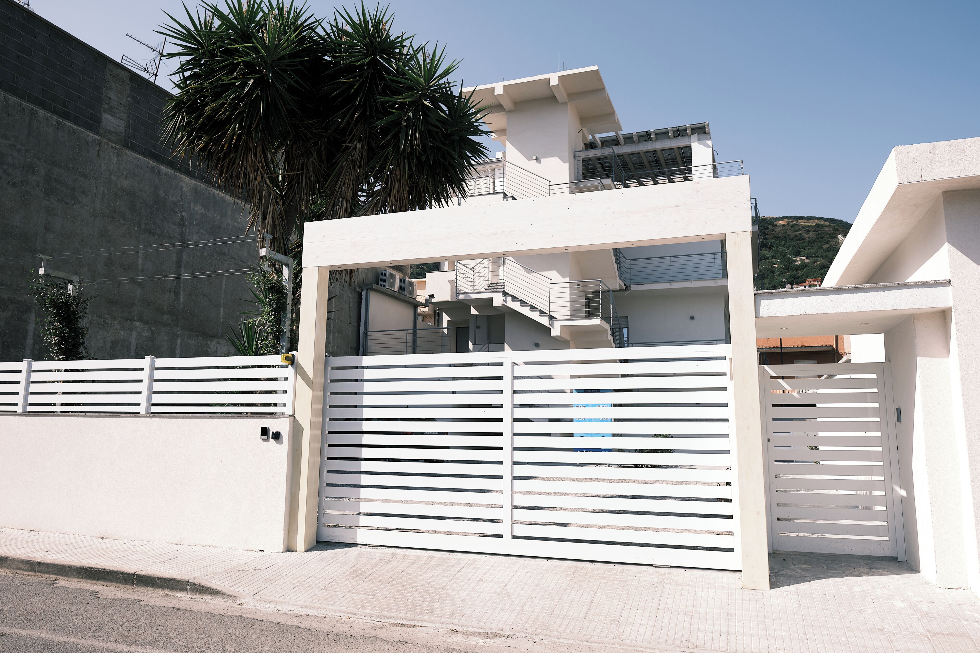A gated entrance to a modern building with a flat roof, a visible staircase, and a palm tree.