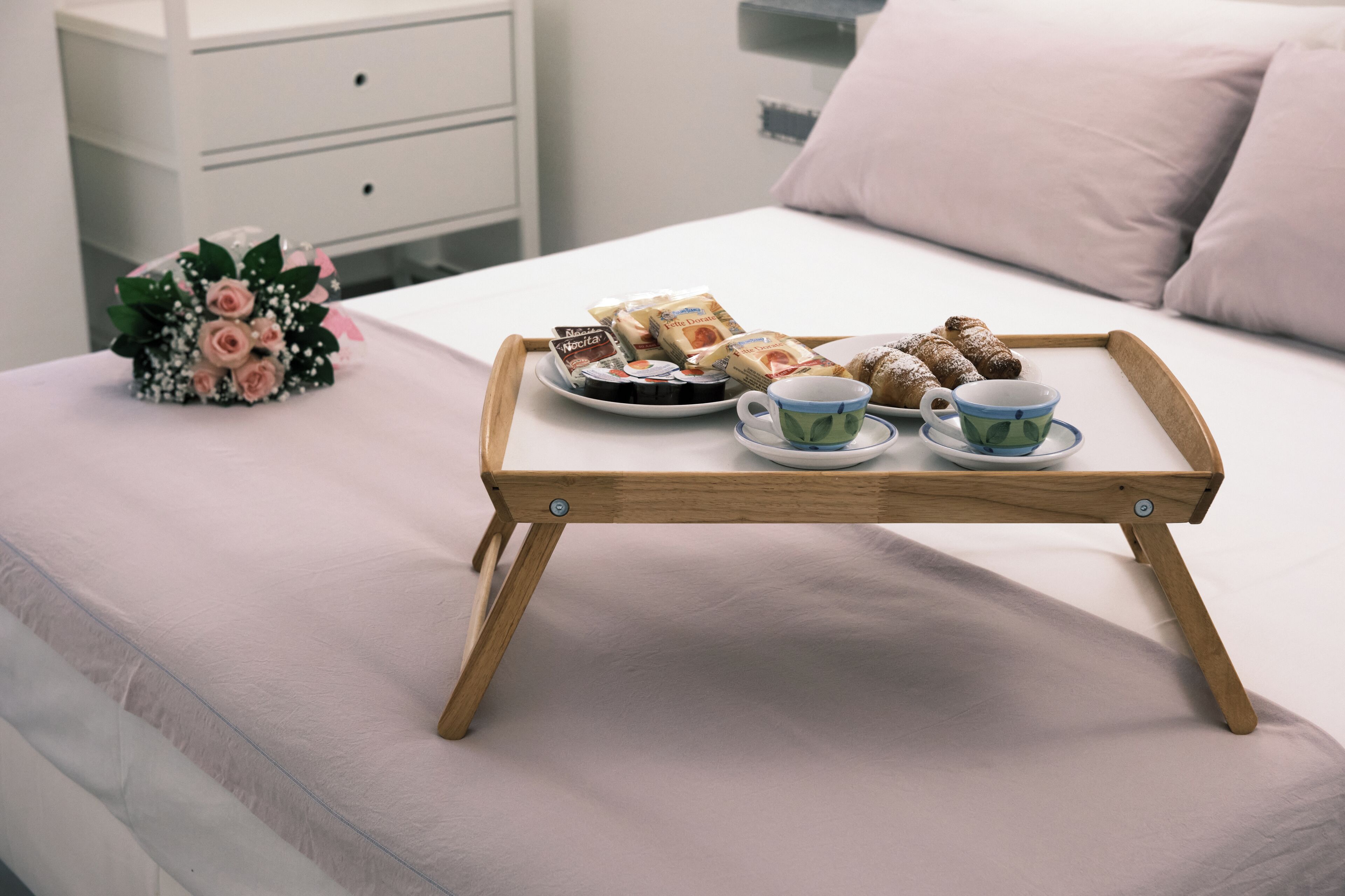 A bed with a wooden tray holding pastries, a cup, and a bouquet of flowers.