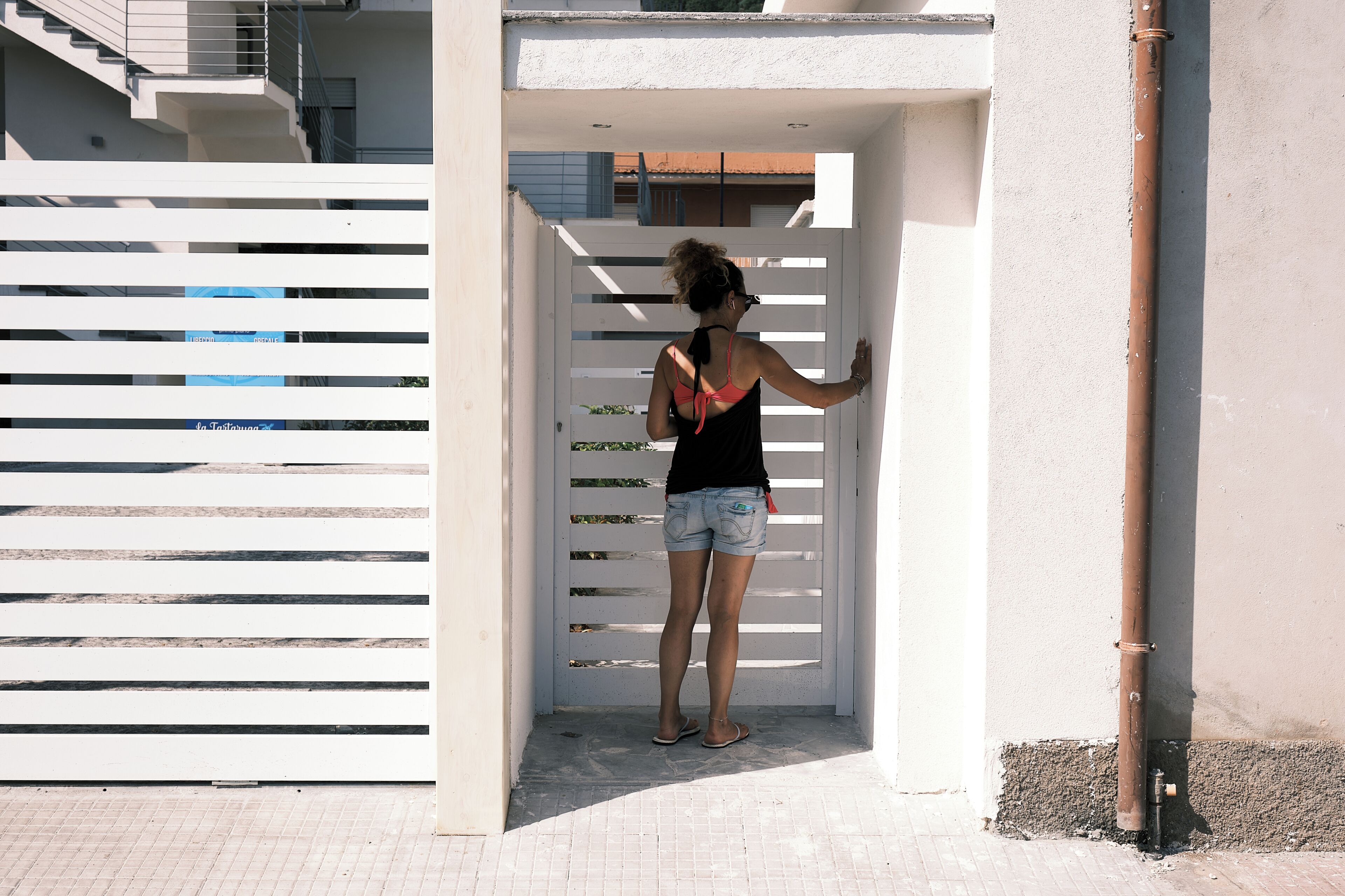 Une personne se tient devant un portail blanc, un bâtiment à la façade blanche, et un tuyau marron est posé sur le sol.