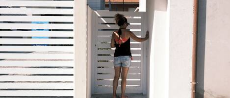 Une personne se tient devant un portail blanc, un bâtiment à la façade blanche, et un tuyau marron est posé sur le sol.