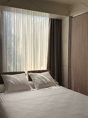 A hotel room with a large bed, two pillows, a window with sheer curtains, and a striped curtain on the side.