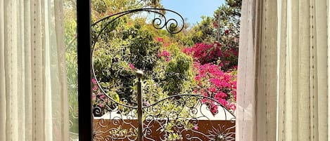 Vista a través de una ventana con cortinas translúcidas, mostrando un jardín con valla de metal decorativa y suelo con diseño.
