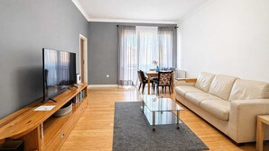 A modern living room with a flat-screen TV, wooden entertainment unit, a glass coffee table, a beige sofa, and a dining area with a table and chairs. - City View Apartment near Nowy Świat (Warsaw)