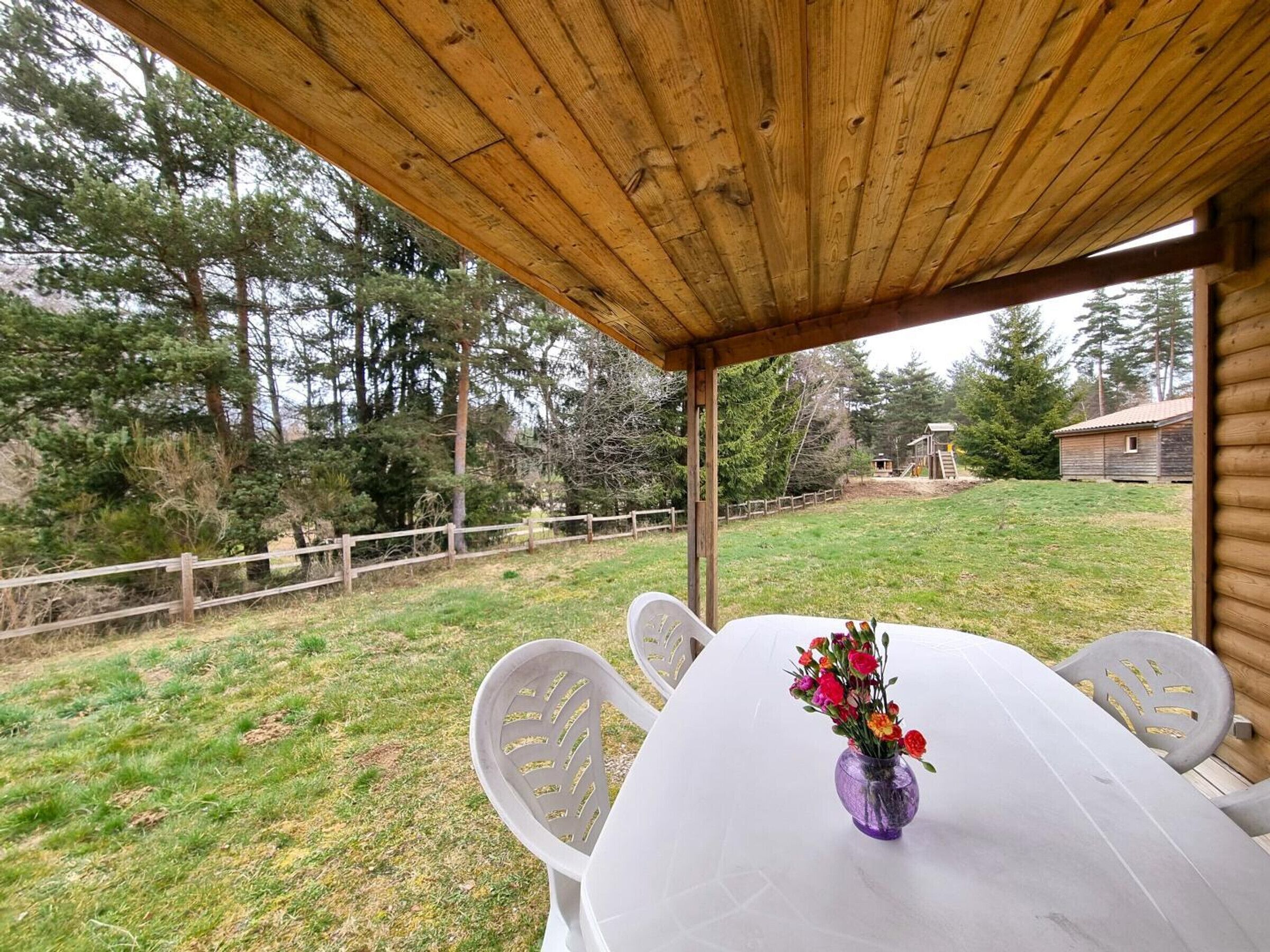 Un área de comedor al aire libre cubierta, con una mesa blanca, un jarrón púrpura con flores y sillas blancas.