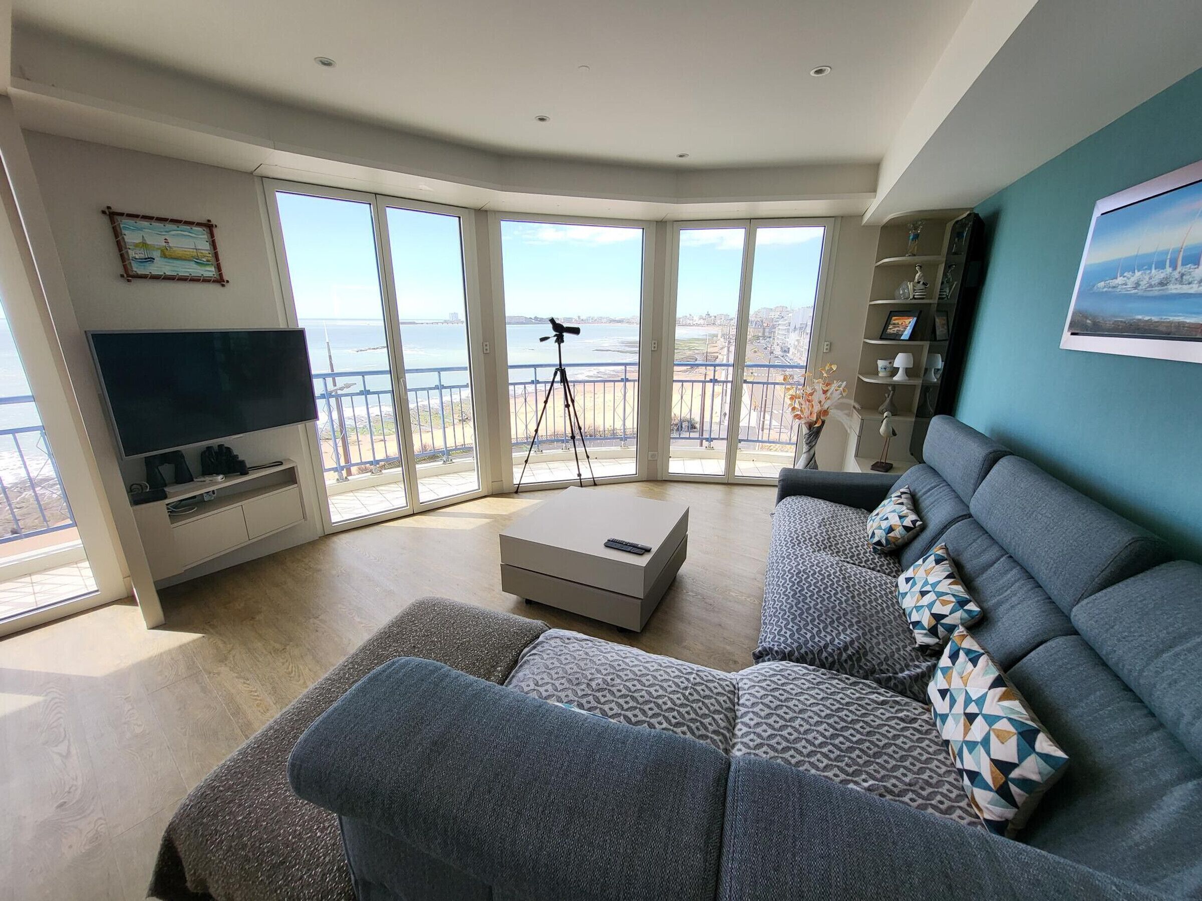 A modern living room with a large TV, a sofa, a coffee table, and a balcony with a view of the sea.