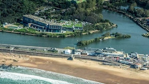 Vista aérea de una zona costera con playa, puerto deportivo y un gran complejo de edificios con varias estructuras.