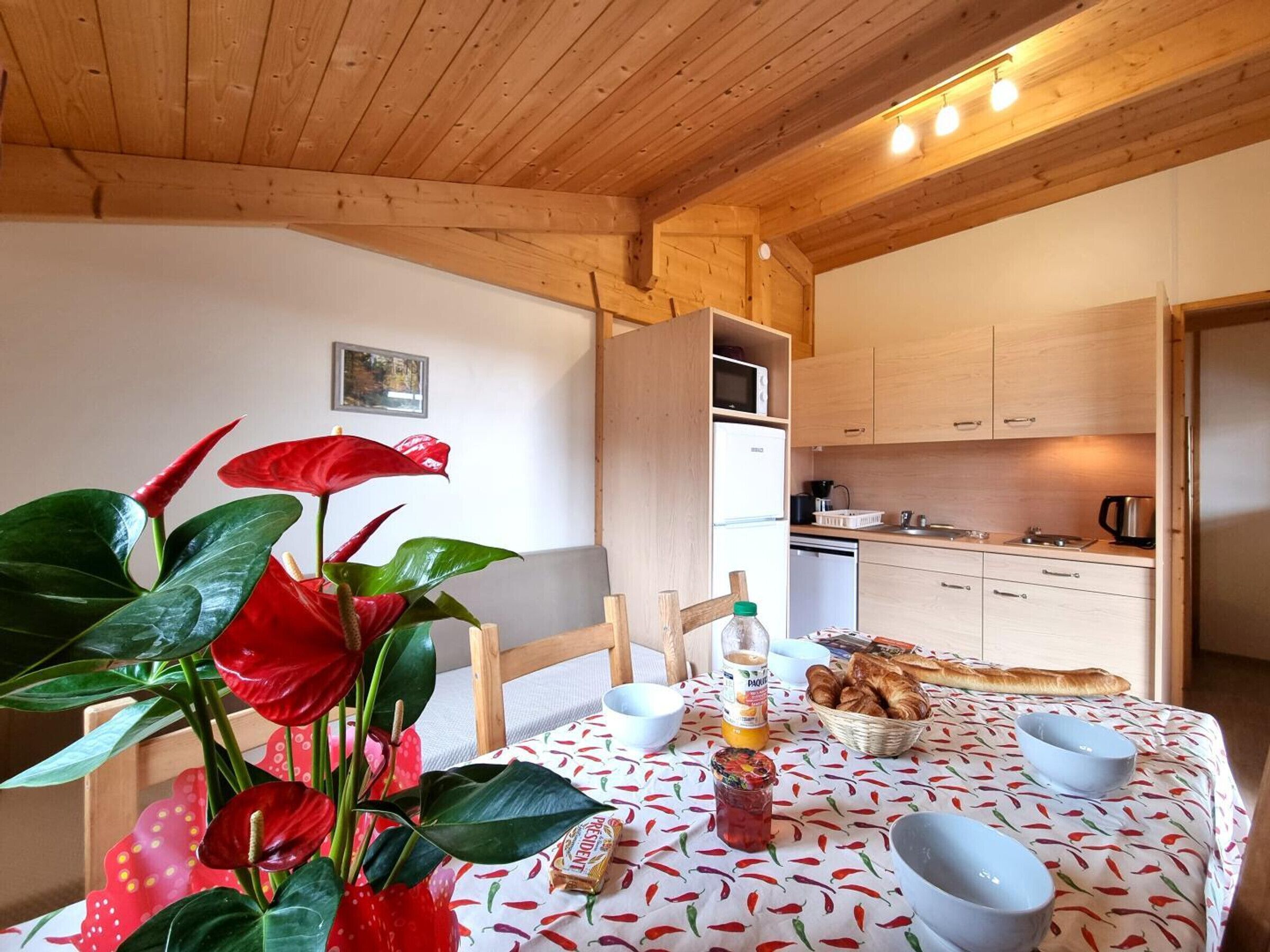 A cozy kitchen with wooden cabinets, a dining table set with a red flower centerpiece, and a breakfast spread.