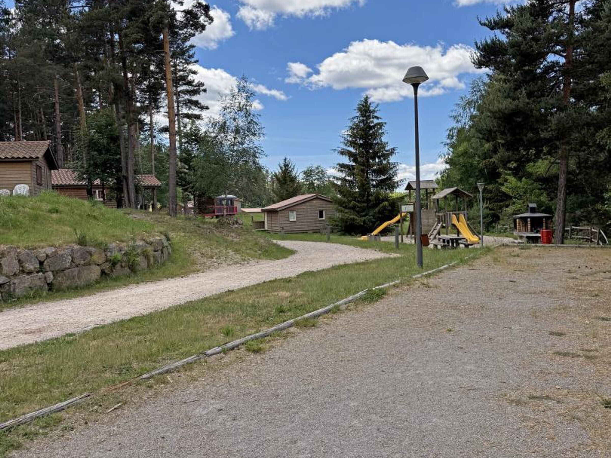 Un camino de grava que lleva a un parque infantil con tobogán y estructuras para escalar, rodeado de cabañas de madera y altos árboles.