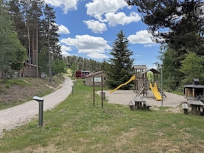 Un parque infantil con tobogán y estructuras para escalar, rodeado de árboles y un camino de grava.