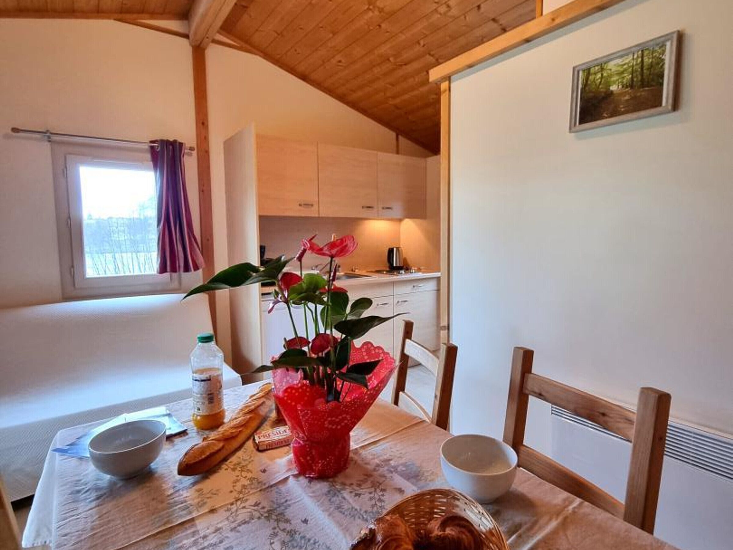 Un comedor con una mesa dispuesta para dos personas, una ventana con cortinas y una cocina al fondo.