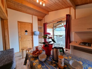 A cozy room with a wooden ceiling, a dining table with a floral tablecloth, a red vase with flowers, a kettle, a bottle of orange juice, and a bowl. - PMR Chalet, pets welcome - 4 guests (Usson-en-Forez)