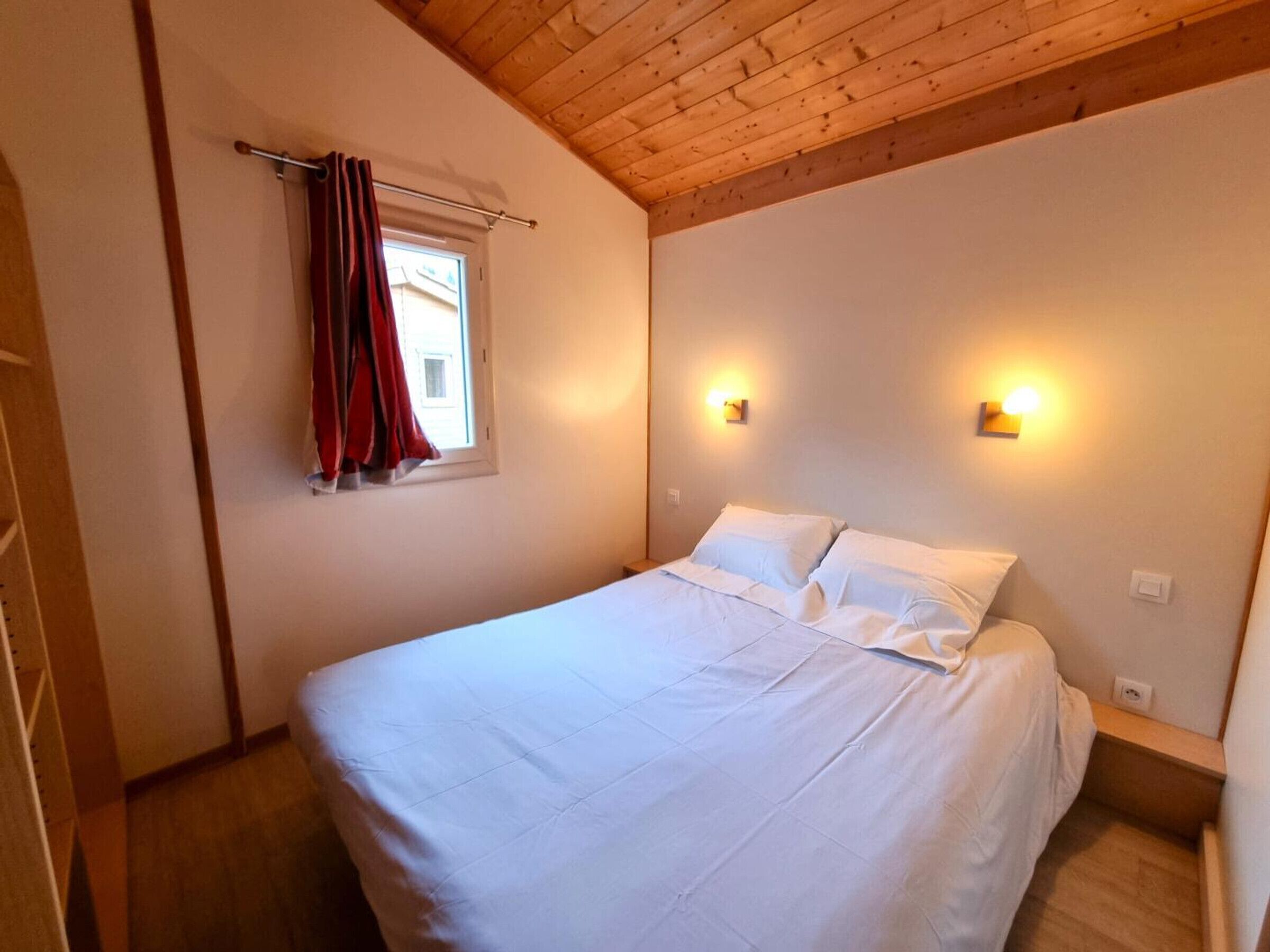 A bedroom with a wooden ceiling, a bed with white bedding, two wall-mounted lights, and a window with red curtains.
