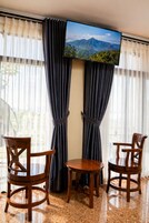 Una habitación con dos sillas de madera, una mesita pequeña, un televisor montado en la pared y cortinas de gasa.