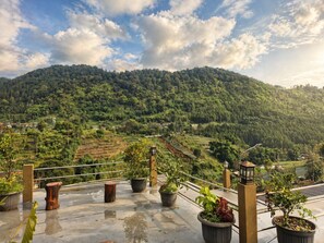 A rooftop terrace with potted plants, a railing, and a view of a lush green valley with terraced fields. - The Green County Homestay (Lembang)